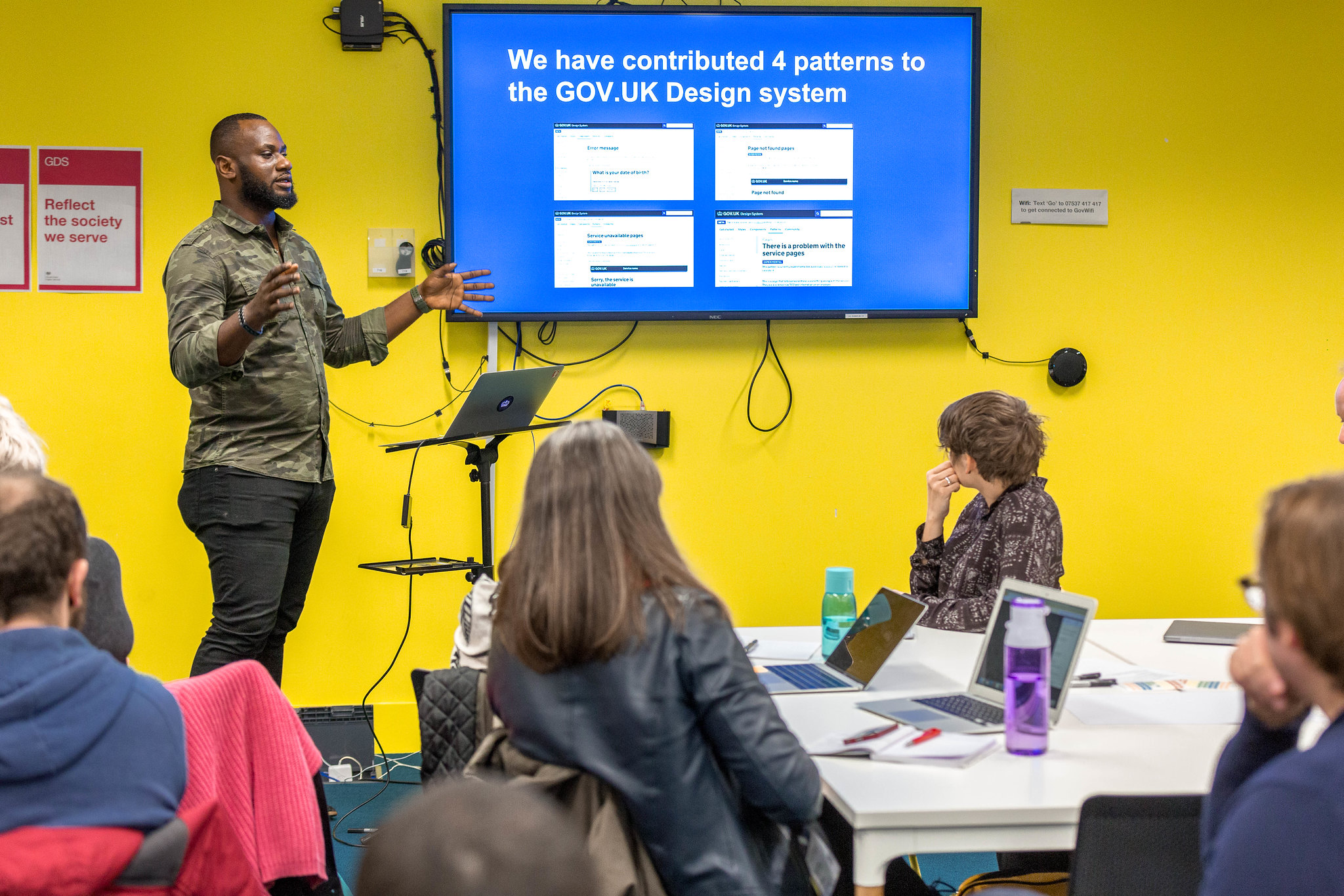 Image of a person presenting to an audience. Next to him is a screen with their presentation and it read: We have contributed 4 patterns to the GOV.UK Design System