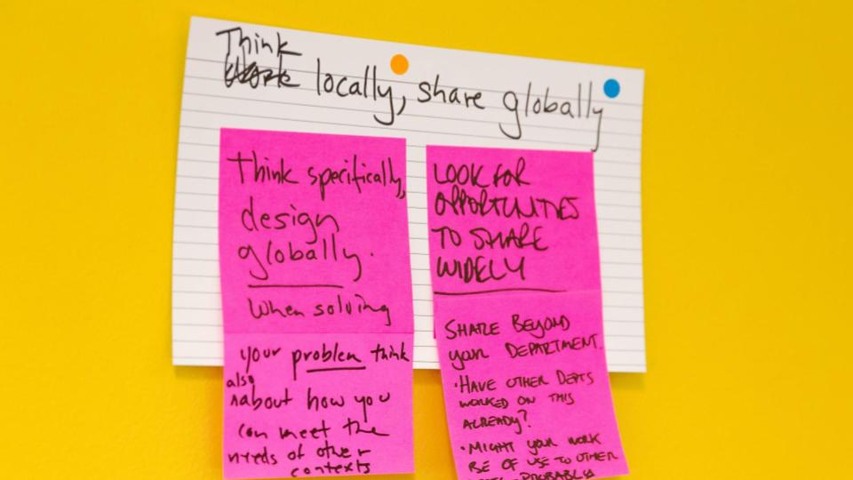 An image of an index card and post it notes stuck onto a wall. It's referring to a co-designed principle for the design system community. It reads "Think locally, share globally"